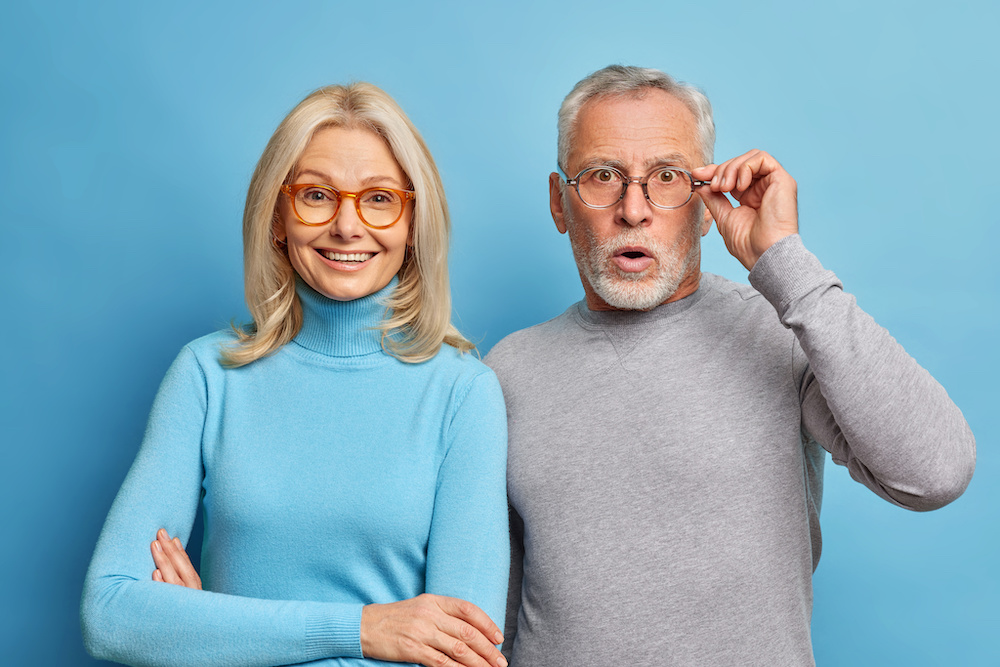 Cheerful middle aged woman and her husband pensioner reacts on shocking news keeps hand on spectacles isolated over blue background. Family age and emotions concept. Senior couple at pose home